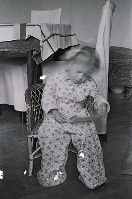 A young child, blurred with motion, sits in a wicker chair holding a small paper or booklet. The scene features a patterned b...