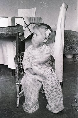 A young girl sits in a wicker chair, wearing a floral patterned robe with a matching headband. The room features a bed with a...