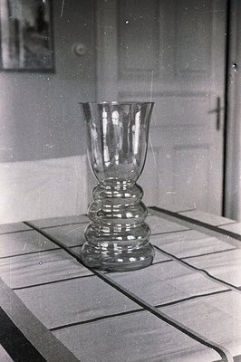 Elegant, stemmed glass vase centered on a textured table. Likely a domestic interior, evident by the closed door and framed a...