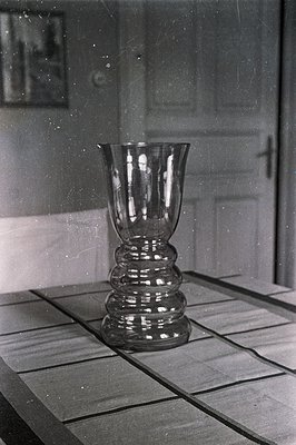 A tall, intricately-designed glass vase stands on a tiled surface. The vase features a flared top and a stepped, bulbous body...