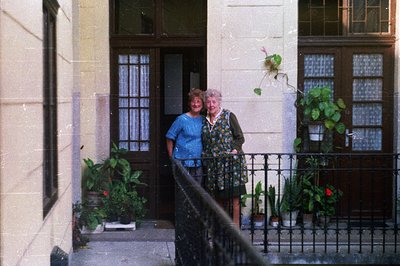 Two women stand on a balcony overlooking a traditional facade. Lush potted plants decorate the wrought-iron railing. The arch...