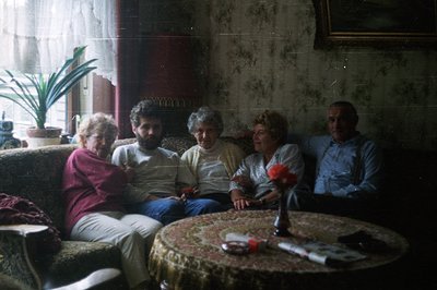 Four individuals seated on a floral-upholstered sofa in a vintage interior. A round table covered with an ornate cloth sits c...