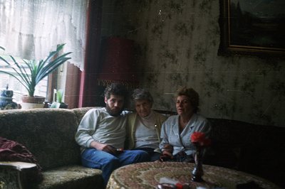 A family scene: a bearded man, older woman, and a younger woman sit on an ornate, floral-patterned sofa in a dimly lit room. ...