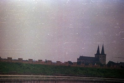 A view of a medieval castle with prominent twin towers, set atop a grassy hill. The castle is backed by a stone wall section....