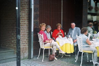 Four adults seated at a table covered with a lace tablecloth in what appears to be an outdoor cafe or restaurant. A potted pl...