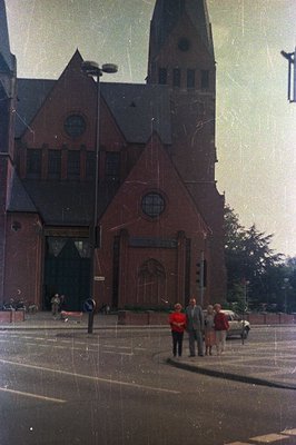 A striking architectural view of a brick church with a tall, pointed spire. Three formally dressed figures – a man in a suit,...