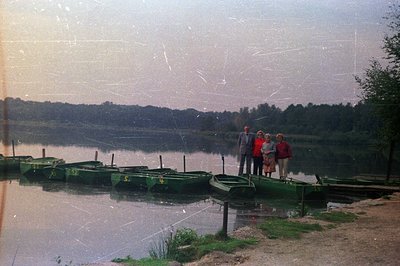 A group of three individuals stands along the shore of a lake, alongside a row of green rental boats. The scene evokes a leis...