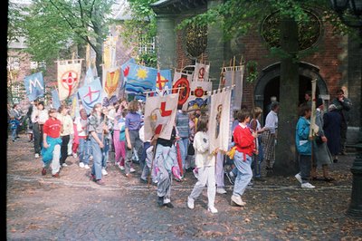 A group of children and adults march through a cobbled square, holding decorated banners—possibly a parade or festival. The a...