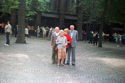 A family portrait: three older individuals stand on a cobblestone plaza, likely a memorial site. Two men flank a woman; all w...