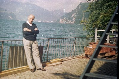 A gentleman in a cable-knit sweater and trousers poses near a lakeside terrace. A stacked pile of bricks is visible, suggesti...