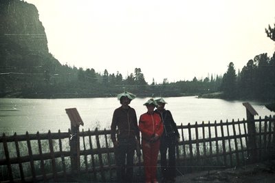 Three figures stand behind a weathered wooden fence overlooking a still, overcast lake backed by a forested hillside. All thr...