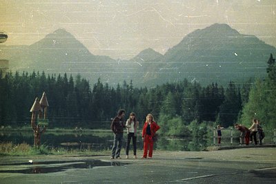 A group of young people stand on a paved road, framed by a dense evergreen forest and towering mountains. The composition sug...