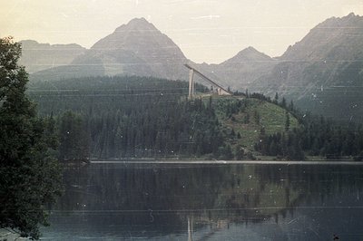 Still lake reflects a towering ski jump structure built on a steep, forested hillside. Dramatic peaks rise in the background,...
