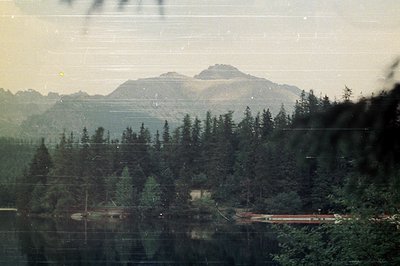 A serene lakeside view showcases a densely forested shoreline, reflecting a towering, snow-capped mountain range. The image l...