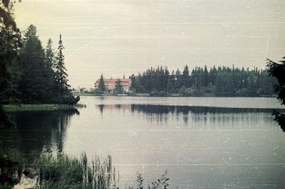 Still water reflects a lakeside building with red tile roof, framed by dense evergreen forest. Likely a resort or institution...
