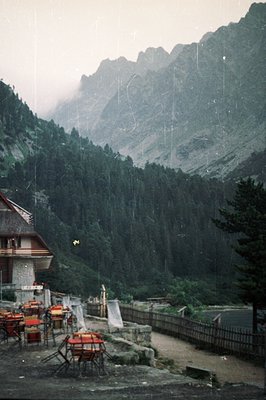 A quaint chalet terrace overlooks a dense forest and steep, rocky mountains shrouded in mist. Rain falls visibly. Red chairs ...
