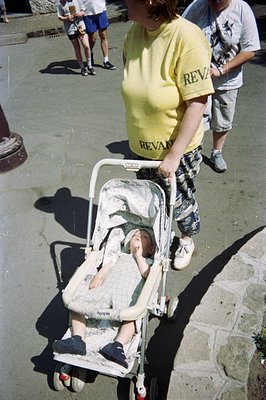 A young child sits in a pale yellow stroller with a patterned fabric cover, appearing contemplative. A man in a yellow tee an...