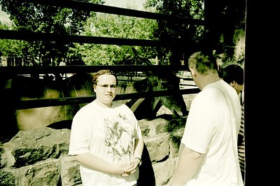 A woman in a graphic tee stands facing a stone wall with a fenced enclosure visible beyond. A man in a white shirt and a part...