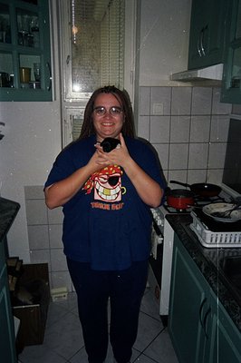 A young man with long hair stands in a kitchen, wearing a dark blue t-shirt featuring a pirate graphic and dark pants. The ki...