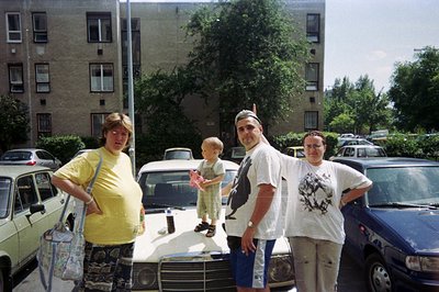 A family of four poses near a dark blue car, framed by a large apartment building. The pregnant woman wears a floral skirt an...