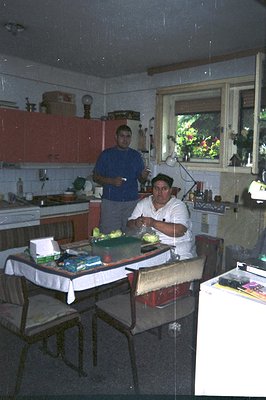 A home kitchen scene, two men preparing food at a table draped with a tablecloth. Coral-colored cabinets, vintage appliances,...