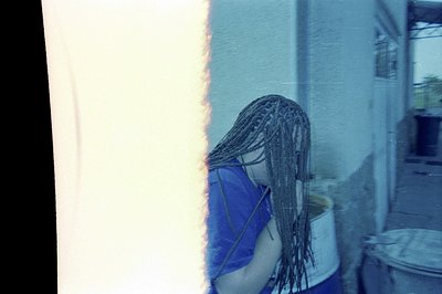 A person, viewed from behind, wears long, dark braided hair and a blue garment, standing near a bucket. The scene appears to ...