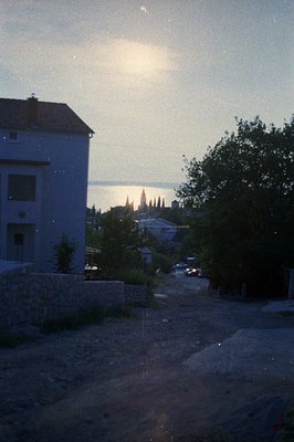 Exterior view of a coastal town at dusk, featuring a two-story white building, stone wall, lush foliage, and a glimpse of a d...