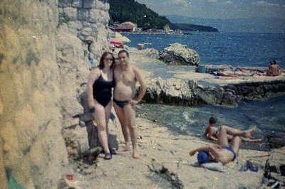 A 1970s snapshot depicts a couple posing near a rocky coastline and beach. The woman wears a black one-piece swimsuit, while ...