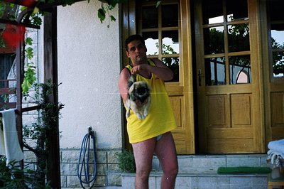 A man in a yellow tank top and no pants holds a pug dog on a small stoop in front of a paneled wooden door. The exterior wall...