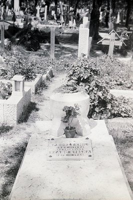 Monochrome image depicts a grave marker in a densely-packed cemetery. The flat stone memorial displays Cyrillic script, likel...