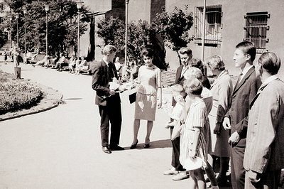 Group of formally dressed young adults and children stand on a paved path lined with greenery and benches. An older couple si...