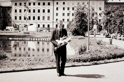A formally dressed man, holding flowers, stands near a pond reflecting a grand, multi-windowed building. Likely 1960s-70s, po...