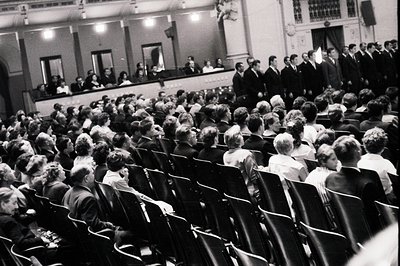 A large audience fills tiered seating, watching a male choral group performing on a raised platform. Formal attire and hairst...