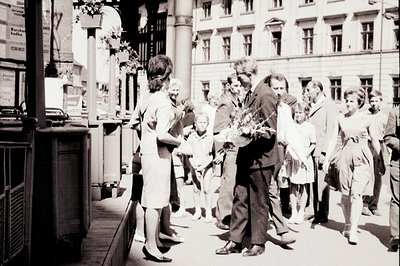 A candid street scene captures a man with flowers interacting with a woman and group of onlookers. Architectural details sugg...