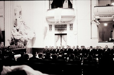 A formal gathering within a grand hall, likely a religious ceremony or official event. A tiered balcony, ornate architectural...