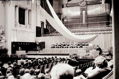 Formal ceremony captured in a grand hall, featuring a stage with seated dignitaries beneath a draped fabric panel. Audience m...