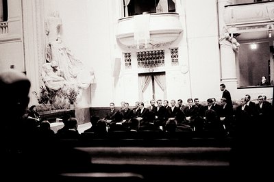 A formal gathering within an ornate building, likely a theater or hall. Numerous men in dark suits are seated in tiered rows....