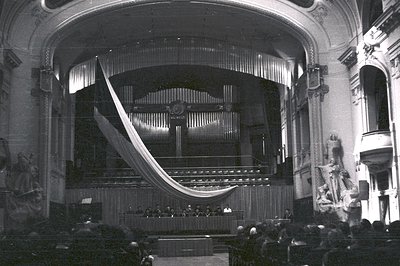 Ornate proscenium stage with a large, draped curtain. Visible audience seated in tiered balconies. Statuary flanks the sides ...