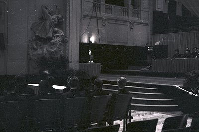 Formal portrait of a group of people observing an elevated stage with a speaker and orchestra. Detailed stone sculpture visib...