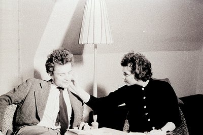 A couple sits closely on upholstered chairs, papers between them. The man, wearing a patterned suit and tie, rests his head o...