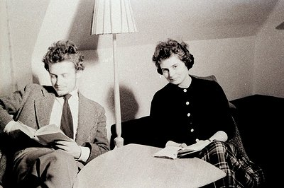 Two figures, a man and a woman, are seated reading books in a warmly lit interior. The man, in a checked blazer and tie, lean...