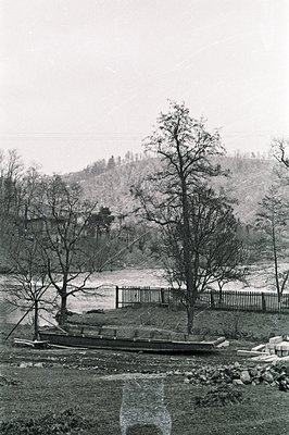 A rustic scene depicts a wooden boat resting on grassy ground near a waterway, framed by bare trees and a weathered wooden fe...