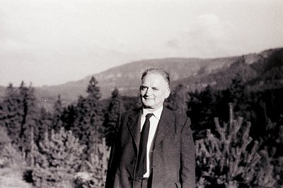 A portrait of a formally dressed man standing amongst evergreen trees, likely in a mountain setting. He wears a dark suit, ti...