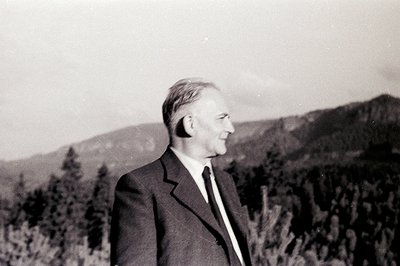A man in a suit and tie stands in profile, gazing towards a mountainous landscape. Appears to be a candid portrait, likely ta...