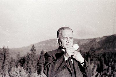 A formally dressed man, likely a priest, holds a host during what appears to be an outdoor religious ceremony. Coniferous tre...