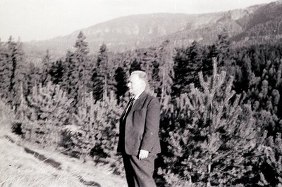 A man in a suit and tie poses beside a densely wooded hillside, likely a forestry area. The landscape suggests an alpine or m...