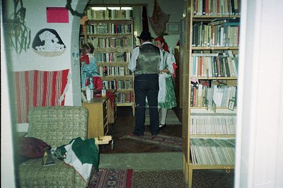 A man in a dark suit and vest stands facing a woman in traditional folk dress in a room filled with bookshelves. The woman we...