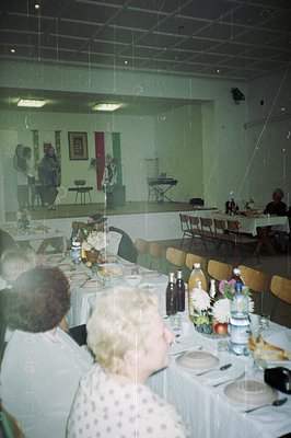 A banquet hall interior; tables draped in white linens are set for a formal dinner. A stage backdrop displays a banner and fl...