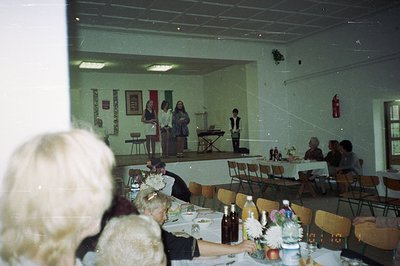 Indoor scene of a gathering, likely a community event or performance. A small stage features three figures; two women and a b...
