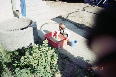 A young boy sits in a red plastic bin, playing with sand near a concrete structure (possibly a well or fountain) and a low st...